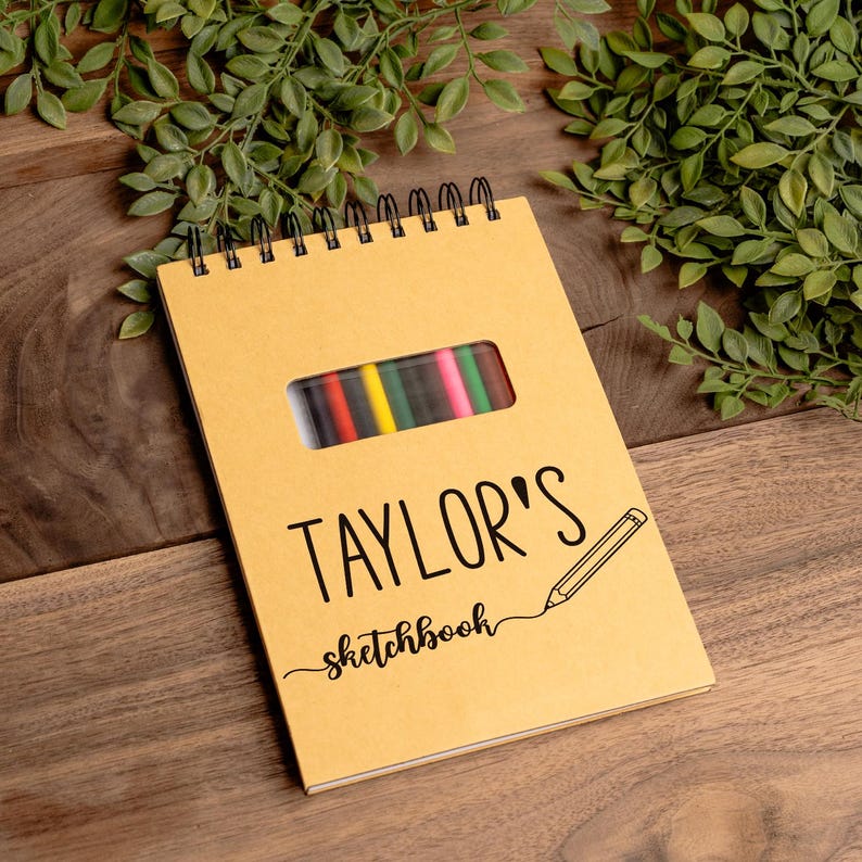 A personalized spiral-bound sketchbook engraved with the name “Taylor’s Sketchbook” on a tan cover, displayed on a wooden surface with green leaves for decoration. The cover features a pencil graphic and a cut-out window revealing 12 colorful pencils inside, stored in an attached pouch. This custom sketchbook makes a thoughtful and practical gift for kids, teens, students, or artists, combining creativity, organization, and personal style in one unique art journal.