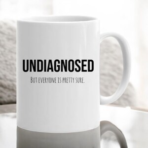May include: White ceramic coffee mug with black text that reads "UNDIAGNOSED BUT EVERYONE IS PRETTY SURE."