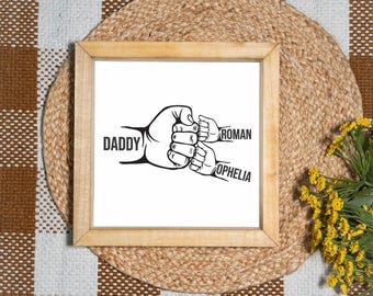 May include: A framed print with a light wood frame. The print features a black and white illustration of three fists bumping. The word "DADDY" is on the left, with "ROMAN" and "OPHELIA" on the right.
