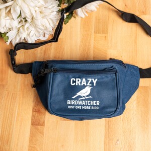 May include: A navy blue fanny pack with the text "CRAZY BIRDWATCHER JUST ONE MORE BIRD" and a white bird graphic. The fanny pack has a black adjustable strap and a zippered pocket. White flowers are in the background.
