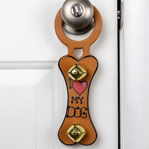 Puede incluir: Un colgador de puerta de cuero marrón en forma de hueso de perro con el texto "Amo a mi perro" y dos campanas doradas. El colgador está sujeto a un pomo de puerta plateado.