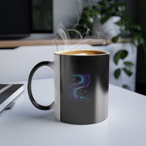 Puede incluir: Una taza de café negra termosensible con un asa curva. El diseño de la taza presenta un colorido patrón de nebulosa arremolinada en tonos de azul, morado y verde azulado. El vapor se eleva del café.