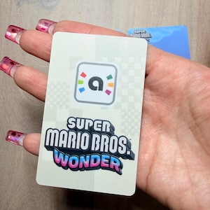 Puede incluir: Una tarjeta rectangular con el logo "Super Mario Bros. Wonder" en dise&ntilde;o 3D y un icono "a" colorido. La tarjeta se sostiene en una mano, con una segunda tarjeta visible en el fondo.
