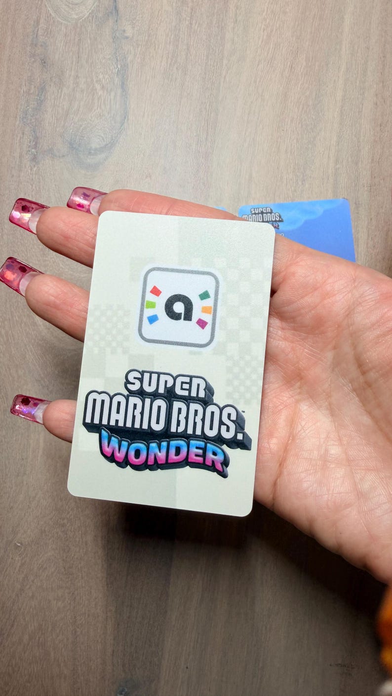 Puede incluir: Una tarjeta rectangular con el logo "Super Mario Bros. Wonder" en dise&ntilde;o 3D y un icono "a" colorido. La tarjeta se sostiene en una mano, con una segunda tarjeta visible en el fondo.