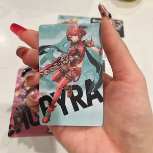 Puede incluir: Una tarjeta rectangular con una ilustraci&oacute;n de personaje. El personaje est&aacute; en una pose din&aacute;mica, vistiendo un atuendo rojo y dorado. El nombre "PYRA" se muestra en letras grandes y en negrita. El fondo es de color azul claro.