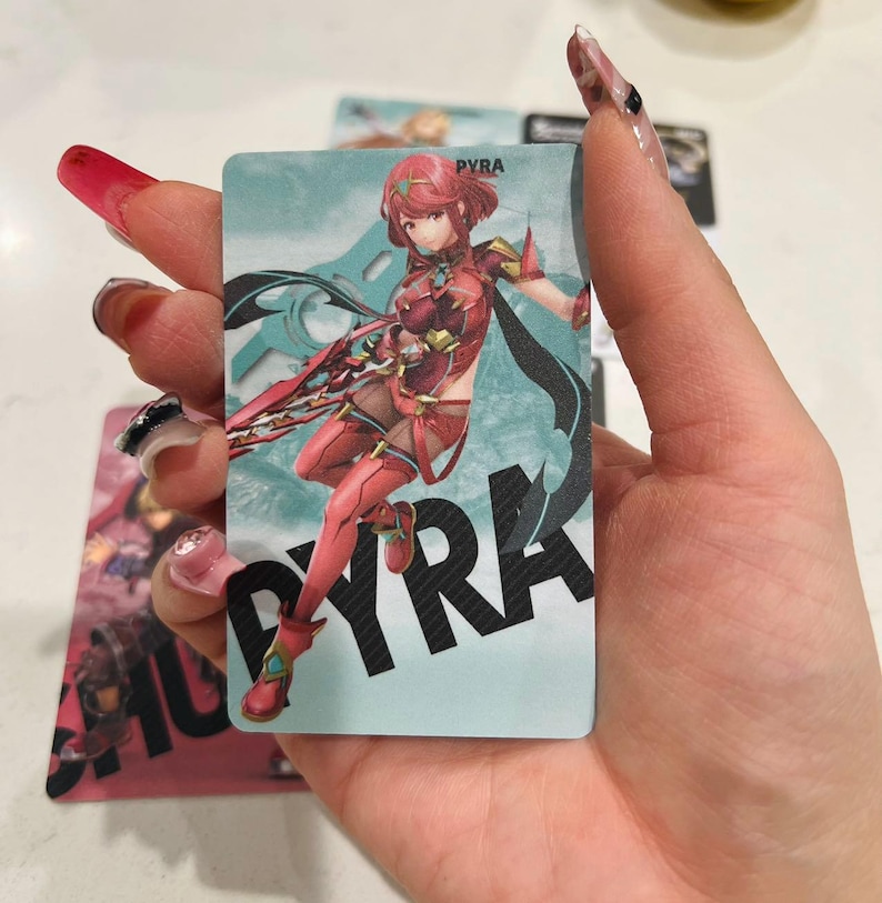 Puede incluir: Una tarjeta rectangular con una ilustraci&oacute;n de personaje. El personaje est&aacute; en una pose din&aacute;mica, vistiendo un atuendo rojo y dorado. El nombre "PYRA" se muestra en letras grandes y en negrita. El fondo es de color azul claro.