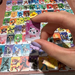 May include: A collection of rectangular trading cards featuring various video game characters arranged in a grid. Each card displays a unique character illustration with a colored background. One card featuring the pink Jigglypuff character is held.