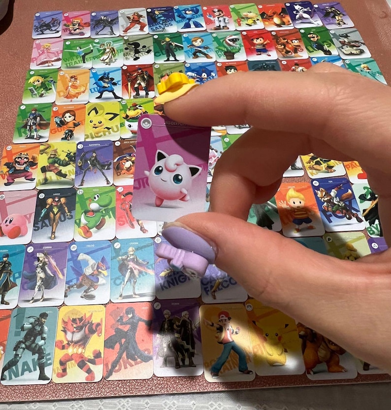 May include: A collection of rectangular trading cards featuring various video game characters arranged in a grid. Each card displays a unique character illustration with a colored background. One card featuring the pink Jigglypuff character is held.