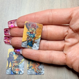 May include: Small rectangular cards featuring colorful illustrations of cartoon characters. The cards are held in a hand with long, pink fingernails. The cards have a silver and gold border.