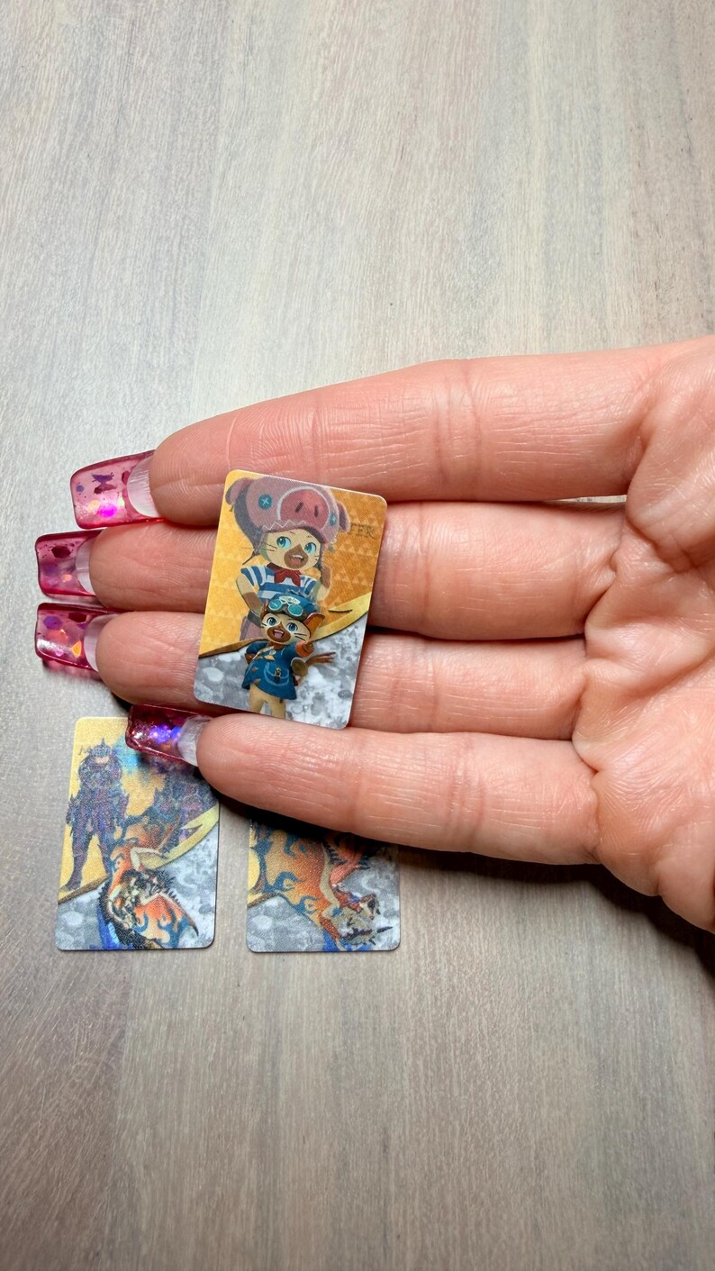 May include: Small rectangular cards featuring colorful illustrations of cartoon characters. The cards are held in a hand with long, pink fingernails. The cards have a silver and gold border.