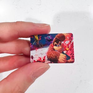 May include: A rectangular sticker depicting Donkey Kong in a dynamic pose. The image shows Donkey Kong against a vibrant background with red and blue hues. The sticker is held by a person. The sticker is a collectible item.