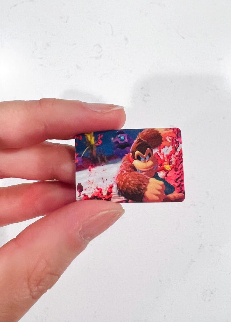 May include: A rectangular sticker depicting Donkey Kong in a dynamic pose. The image shows Donkey Kong against a vibrant background with red and blue hues. The sticker is held by a person. The sticker is a collectible item.