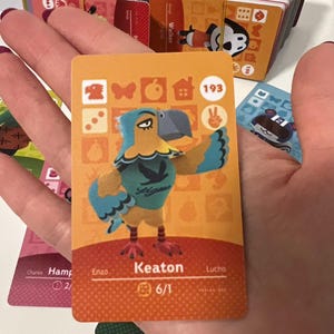 May include: A rectangular trading card featuring a cartoon eagle character named Keaton. The card has an orange background with various icons, the number 193, and the text Keaton 6/1. The character is blue and yellow with a grey beak.