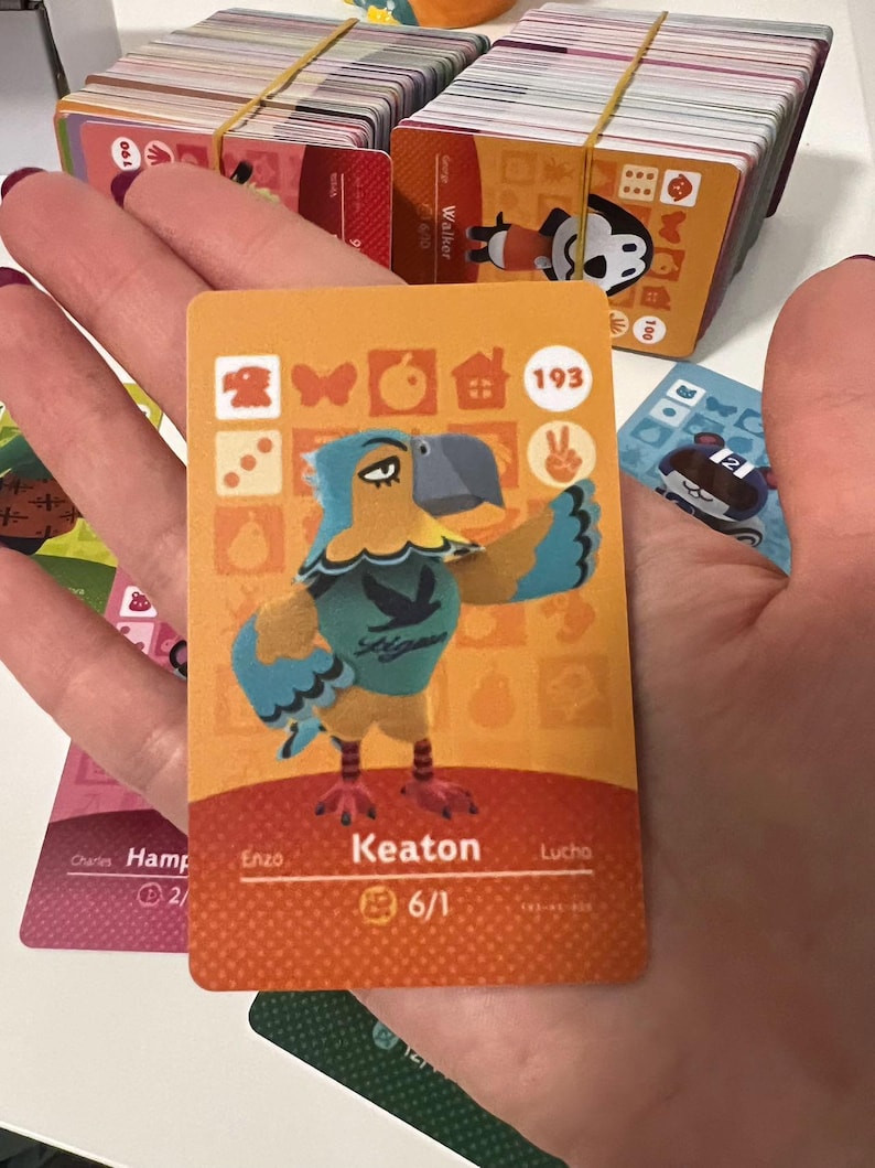May include: A rectangular trading card featuring a cartoon eagle character named Keaton. The card has an orange background with various icons, the number 193, and the text Keaton 6/1. The character is blue and yellow with a grey beak.