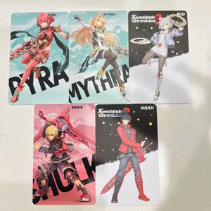 Puede incluir: Cinco tarjetas de personajes con ilustraciones de personajes de Xenoblade Chronicles. Las tarjetas muestran los personajes Pyra, Mythra, Mio, Shulk y Noah. Cada tarjeta tiene el nombre del personaje impreso en letras grandes.