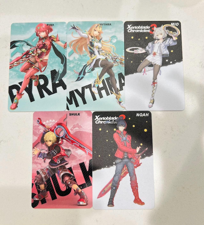 Puede incluir: Cinco tarjetas de personajes con ilustraciones de personajes de Xenoblade Chronicles. Las tarjetas muestran los personajes Pyra, Mythra, Mio, Shulk y Noah. Cada tarjeta tiene el nombre del personaje impreso en letras grandes.