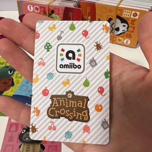 May include: A rectangular Animal Crossing amiibo card featuring a white and grey diagonal striped background with colourful icons of fruit, shells, balloons, and insects. The card displays the amiibo logo and the Animal Crossing game title logo.
