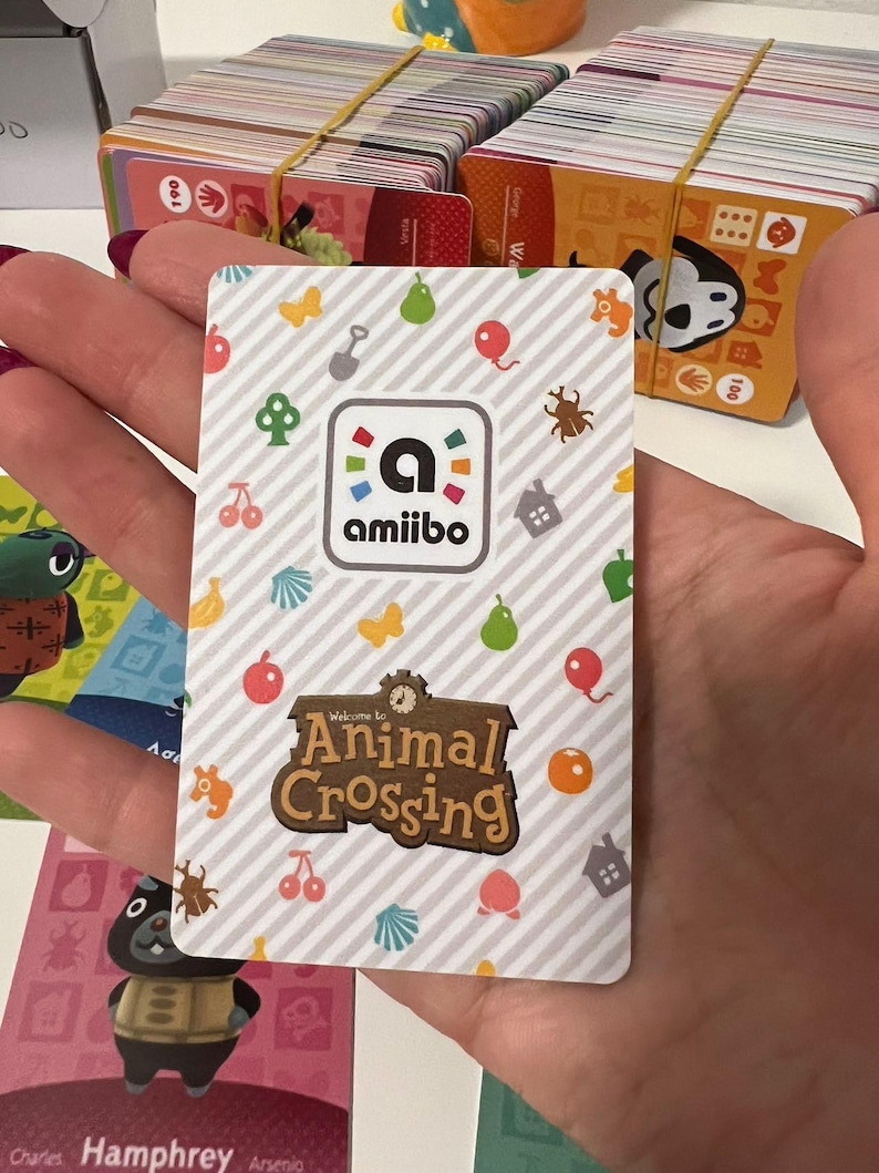 May include: A rectangular Animal Crossing amiibo card featuring a white and grey diagonal striped background with colourful icons of fruit, shells, balloons, and insects. The card displays the amiibo logo and the Animal Crossing game title logo.