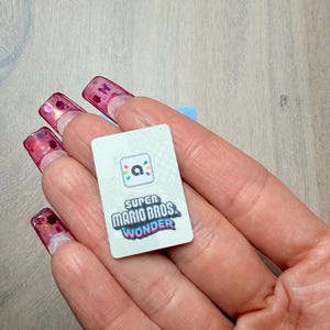 Puede incluir: Una peque&ntilde;a tarjeta rectangular con el logotipo de "Super Mario Bros. Wonder". La tarjeta tiene un fondo blanco con texto y gr&aacute;ficos coloridos. La tarjeta se sostiene en una mano con u&ntilde;as largas y rosas.