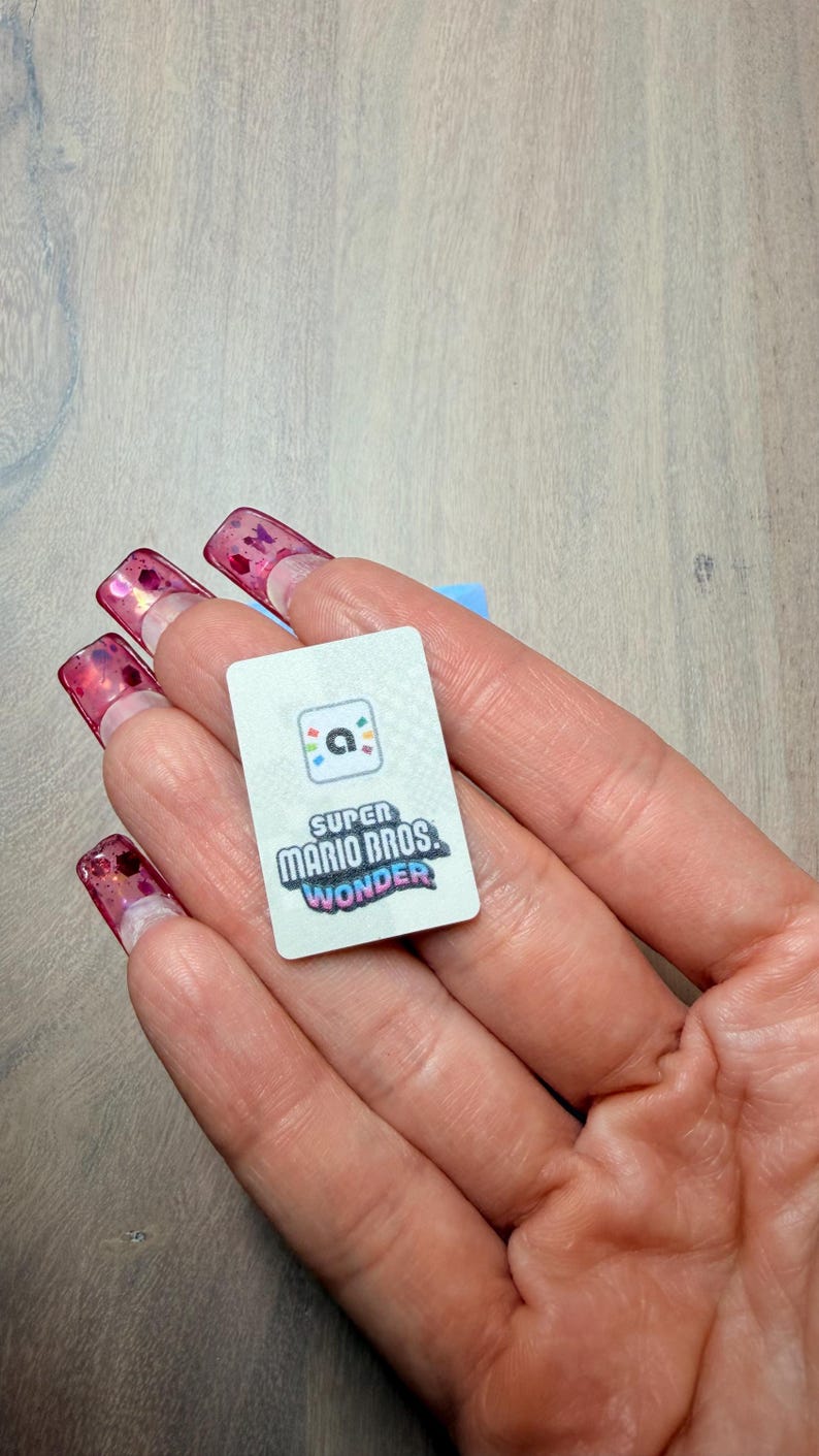 Puede incluir: Una peque&ntilde;a tarjeta rectangular con el logotipo de "Super Mario Bros. Wonder". La tarjeta tiene un fondo blanco con texto y gr&aacute;ficos coloridos. La tarjeta se sostiene en una mano con u&ntilde;as largas y rosas.