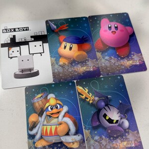 Op de afbeelding: Vijf rechthoekige verzamelkaarten met videogamepersonages. E&eacute;n kaart is wit met de tekst BOX BOY! en een illustratie. De andere vier kaarten hebben blauwe sterrenachtergronden en tonen Kirby, Bandana Waddle Dee, King Dedede en Meta Knight.