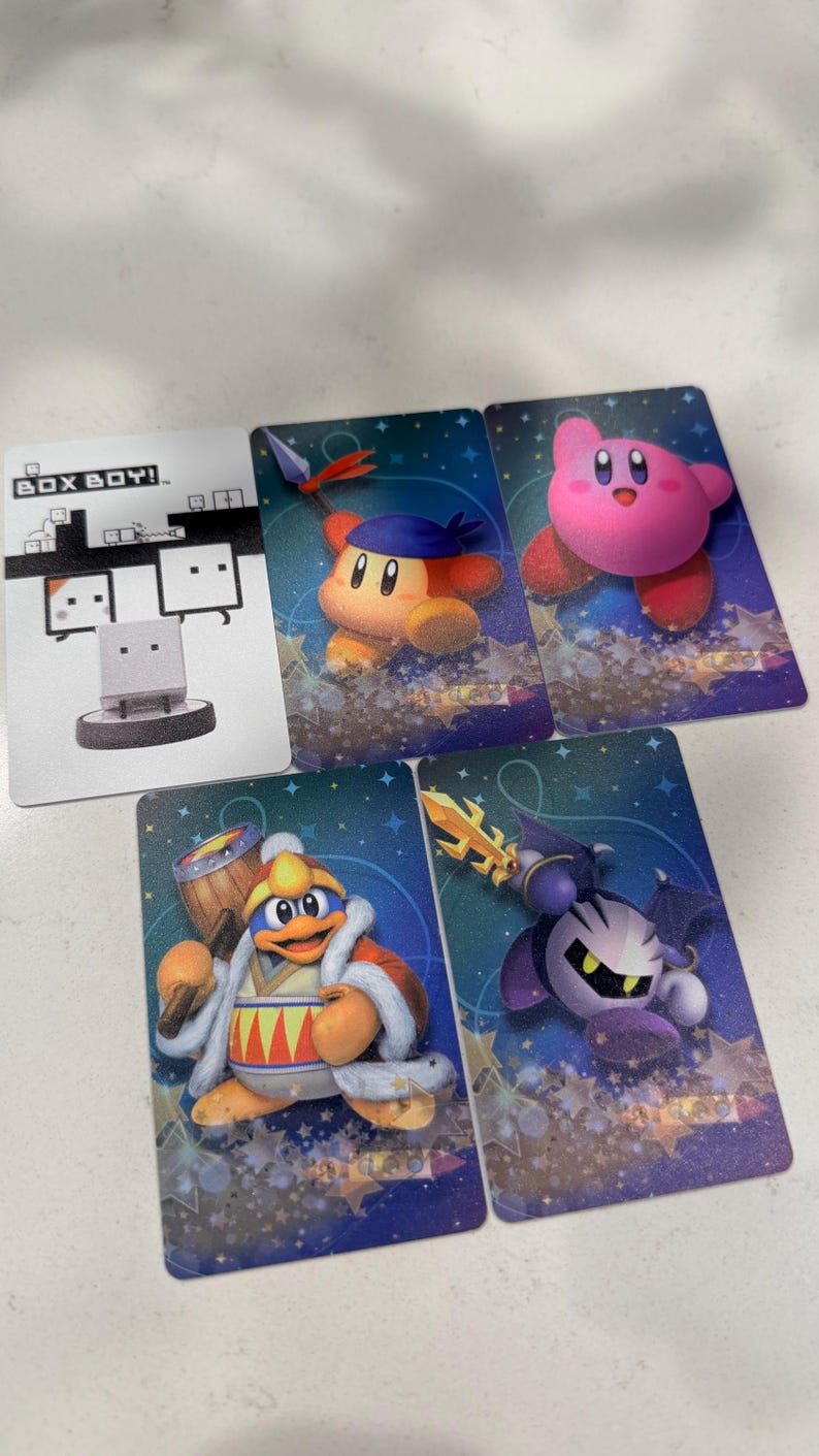 Op de afbeelding: Vijf rechthoekige verzamelkaarten met videogamepersonages. E&eacute;n kaart is wit met de tekst BOX BOY! en een illustratie. De andere vier kaarten hebben blauwe sterrenachtergronden en tonen Kirby, Bandana Waddle Dee, King Dedede en Meta Knight.