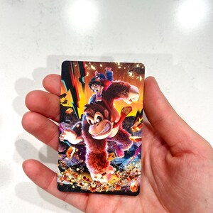 May include: A rectangular card with a colourful illustration of a gorilla-like character with a smaller figure riding on its back. The image has a dynamic composition with vibrant colours and a detailed background.