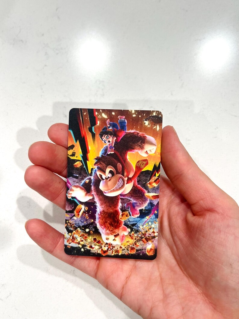 May include: A rectangular card with a colourful illustration of a gorilla-like character with a smaller figure riding on its back. The image has a dynamic composition with vibrant colours and a detailed background.