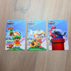 Puede incluir: Tres tarjetas coleccionables de Super Mario Bros. Wonder. Las tarjetas presentan a Poplin & Prince Florian, Captain Toad & Talking Flower y Elephant Mario. Cada tarjeta tiene un fondo azul con ilustraciones de personajes.