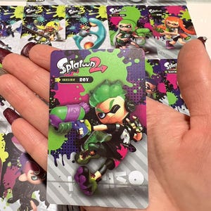 May include: A collection of Splatoon 2 trading cards featuring colorful characters and designs. The central card displays "Inkline Boy" with a green-haired character holding a purple and green weapon. Other cards show various characters and the Splatoon logo.