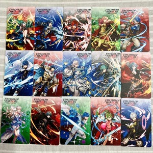 May include: A collection of fifteen rectangular trading cards featuring Fire Emblem Warriors Three Hopes character illustrations. Each card displays a unique anime-style warrior in a dynamic pose with vibrant magical effects, colourful backgrounds, and the Fire Emblem Warriors Three Hopes logo printed at the top.