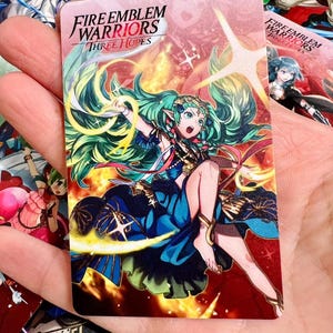 May include: A rectangular trading card featuring the Fire Emblem Warriors Three Hopes logo at the top. The card displays an anime-style character with long green hair, wearing a blue dress, surrounded by yellow light effects and a red background.