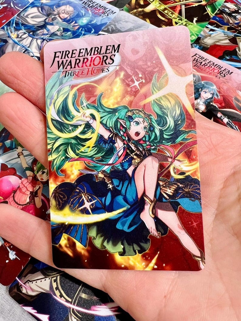May include: A rectangular trading card featuring the Fire Emblem Warriors Three Hopes logo at the top. The card displays an anime-style character with long green hair, wearing a blue dress, surrounded by yellow light effects and a red background.