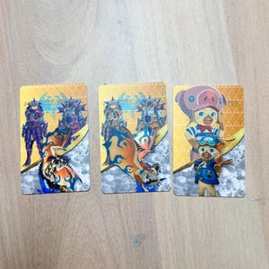 May include: Three rectangular trading cards featuring colorful illustrations of characters and creatures. The cards have a gold and white background with the text "Monster Hunter" visible. The cards are likely part of a collectible card game.