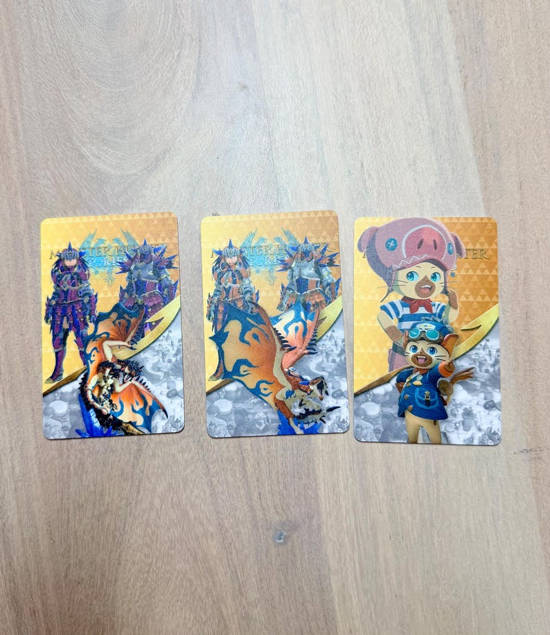 May include: Three rectangular trading cards featuring colorful illustrations of characters and creatures. The cards have a gold and white background with the text "Monster Hunter" visible. The cards are likely part of a collectible card game.