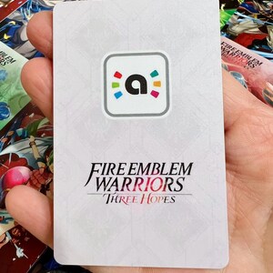 May include: A rectangular white game card featuring a central square icon with a lowercase letter a surrounded by colourful dots. Below the icon, the text reads Fire Emblem Warriors Three Hopes in black and red lettering against a subtle geometric pattern.