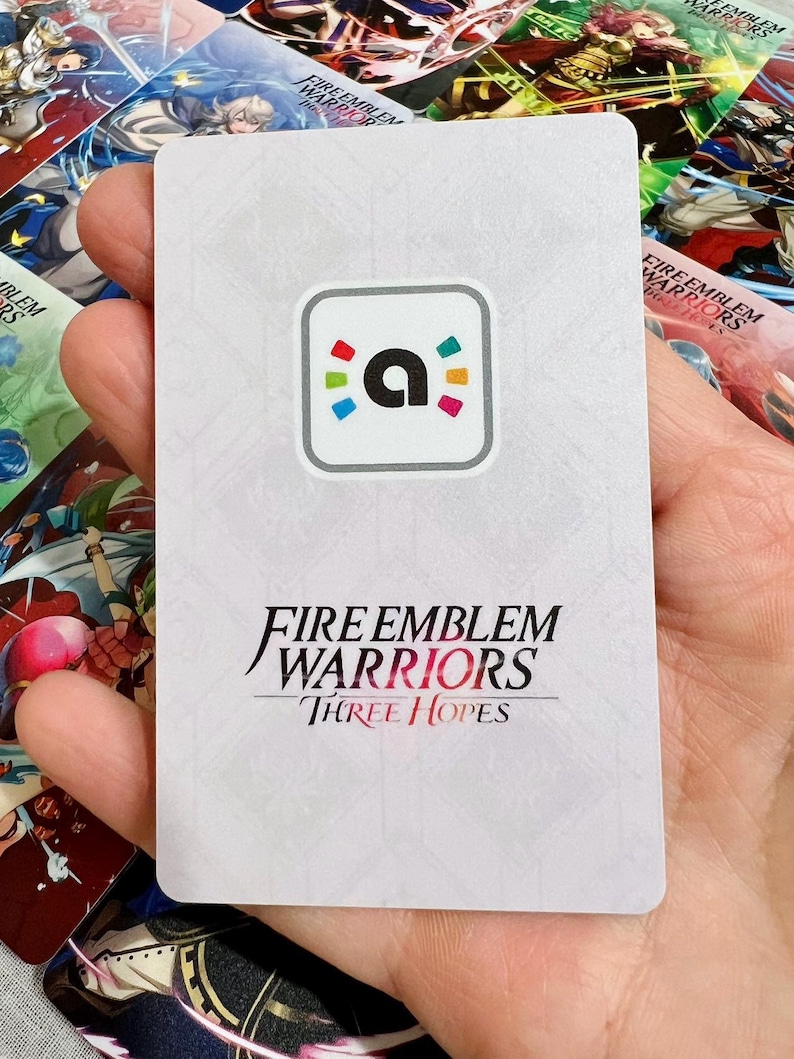 May include: A rectangular white game card featuring a central square icon with a lowercase letter a surrounded by colourful dots. Below the icon, the text reads Fire Emblem Warriors Three Hopes in black and red lettering against a subtle geometric pattern.