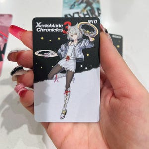 Puede incluir: Una tarjeta coleccionable negra con una ilustraci&oacute;n de anime de un personaje de Xenoblade Chronicles 3. El personaje es una mujer con cabello blanco, vestida con un vestido blanco, chaqueta y medias negras. La tarjeta tiene el texto "Xenoblade Chronicles 3" y "MIO".
