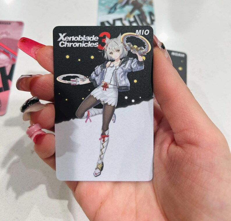 Puede incluir: Una tarjeta coleccionable negra con una ilustraci&oacute;n de anime de un personaje de Xenoblade Chronicles 3. El personaje es una mujer con cabello blanco, vestida con un vestido blanco, chaqueta y medias negras. La tarjeta tiene el texto "Xenoblade Chronicles 3" y "MIO".