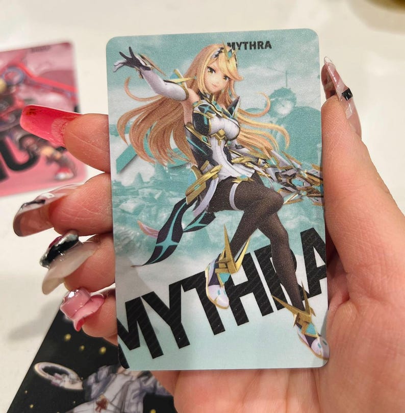 Puede incluir: Tarjeta coleccionable con una ilustraci&oacute;n de estilo anime de un personaje femenino con cabello rubio y un atuendo blanco, negro y dorado. La tarjeta tiene el nombre "MYTHRA" impreso en letras grandes y en negrita.