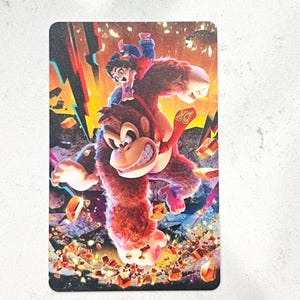 May include: A rectangular card with a colourful illustration of Donkey Kong with a person riding on his back. The artwork has a dynamic, action-packed feel with bright colours, including red, orange, and blue, and a "DK" logo.