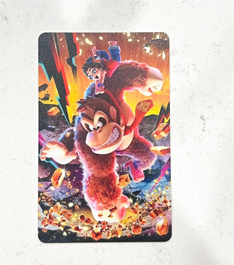 May include: A rectangular card with a colourful illustration of Donkey Kong with a person riding on his back. The artwork has a dynamic, action-packed feel with bright colours, including red, orange, and blue, and a "DK" logo.