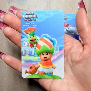 Puede incluir: Una tarjeta coleccionable de Super Mario Bros. Wonder con Poplin y el Pr&iacute;ncipe Florian. La tarjeta muestra una colorida ilustraci&oacute;n de los personajes con un fondo vibrante. La tarjeta se sostiene en una mano.