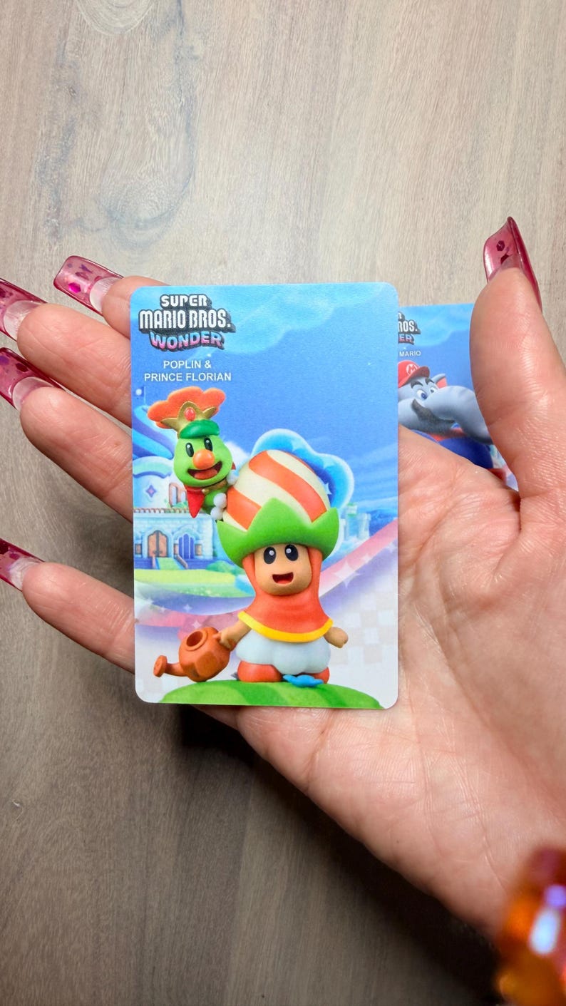 Puede incluir: Una tarjeta coleccionable de Super Mario Bros. Wonder con Poplin y el Pr&iacute;ncipe Florian. La tarjeta muestra una colorida ilustraci&oacute;n de los personajes con un fondo vibrante. La tarjeta se sostiene en una mano.