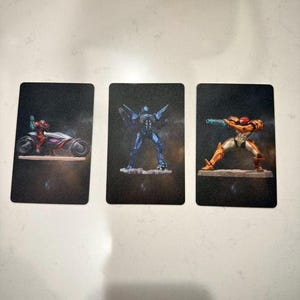 May include: Three trading cards featuring illustrations of characters from a science fiction video game. The cards depict a character on a futuristic motorcycle, a blue robotic figure, and a character in orange and gold armor.