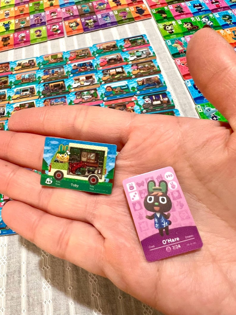 May include: Two rectangular trading cards featuring Animal Crossing characters. One card shows a green camper van with the name Toby, while the other is a pink card featuring a brown rabbit character named O'Hare with the text 7/24 and 390.