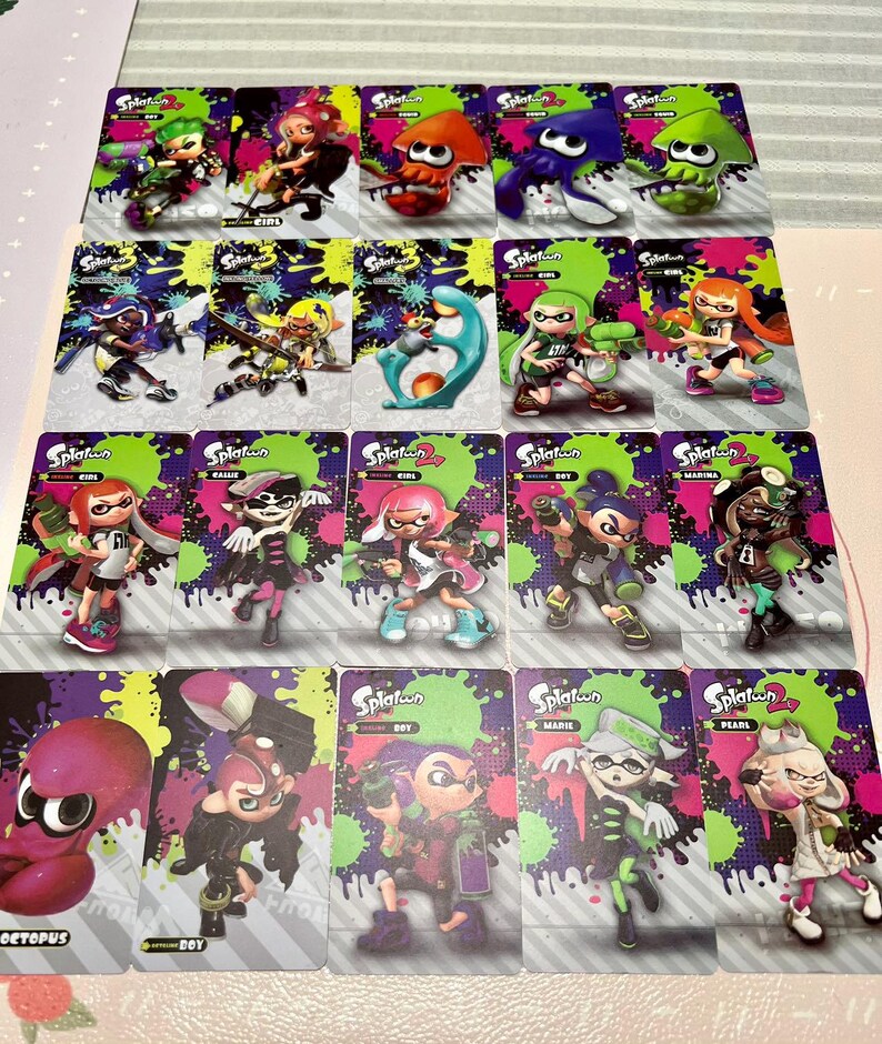 May include: A collection of Splatoon character trading cards. Each card features a different character in vibrant colors, set against a background of green and purple splatters. The cards are arranged in rows and columns.