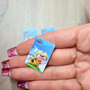 Puede incluir: Una peque&ntilde;a pegatina rectangular con una colorida ilustraci&oacute;n de un personaje del juego Super Mario Bros. Wonder. La pegatina tiene un fondo azul con el logotipo del juego y un personaje de dibujos animados. Se ven otras pegatinas.