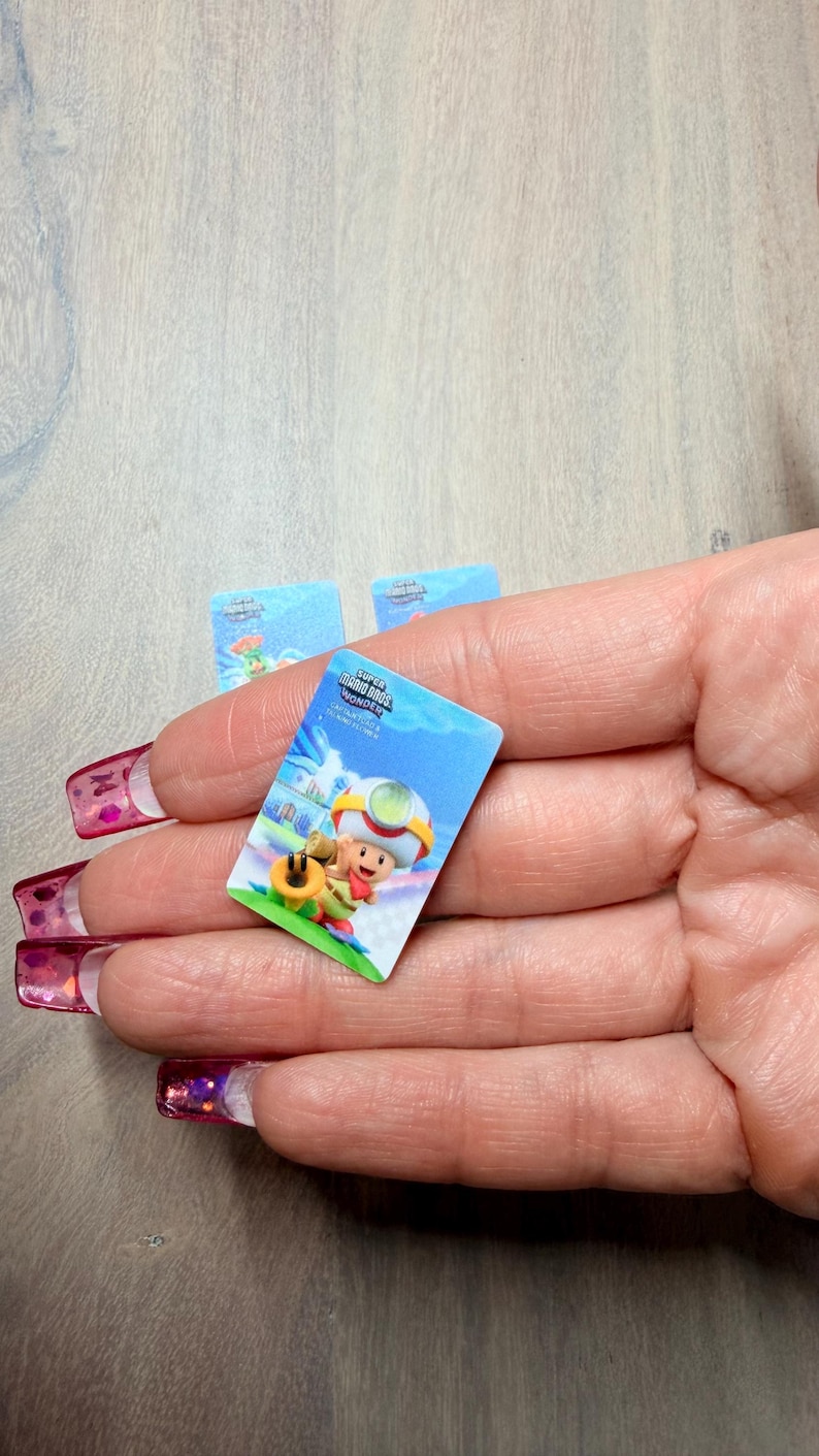 Puede incluir: Una peque&ntilde;a pegatina rectangular con una colorida ilustraci&oacute;n de un personaje del juego Super Mario Bros. Wonder. La pegatina tiene un fondo azul con el logotipo del juego y un personaje de dibujos animados. Se ven otras pegatinas.
