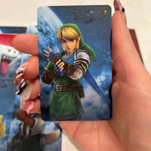 K&ouml;nnte beinhalten: Eine rechteckige Sammelkarte mit einer digitalen Illustration der Figur Link aus der The Legend of Zelda-Reihe, die eine gr&uuml;ne Tunika und M&uuml;tze tr&auml;gt, mit dem Bayonetta-Logo in der oberen rechten Ecke vor einem blauen Weltraumhintergrund.